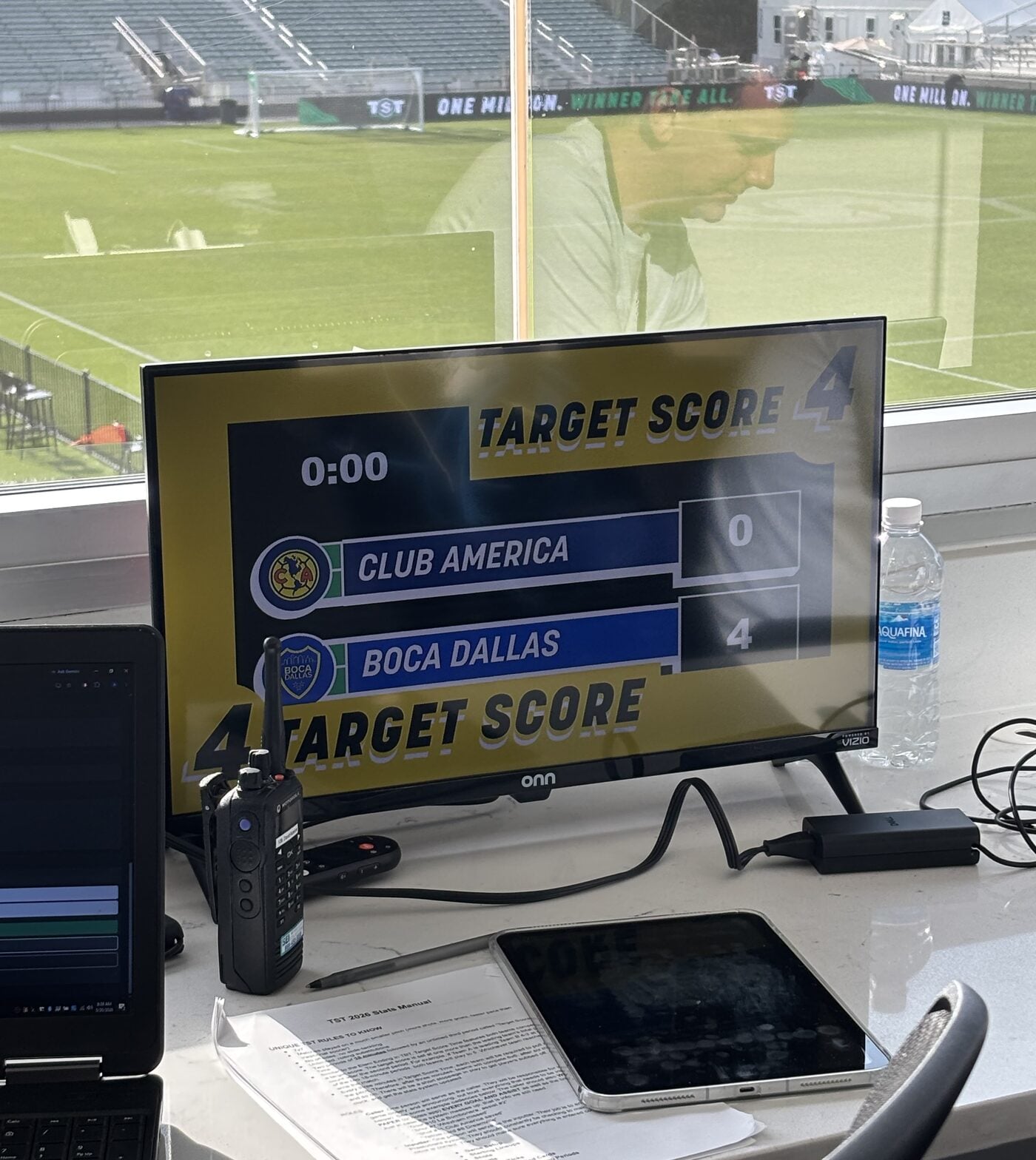 The booth setup at TST: a monitor showing the scoreboard, a tablet for the control panel, and the main stadium pitch beyond the glass